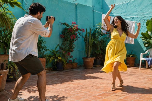 Two adults film a music video in a sunlit courtyard; one dances in a bright yellow dress while the other records on a small handheld camera.